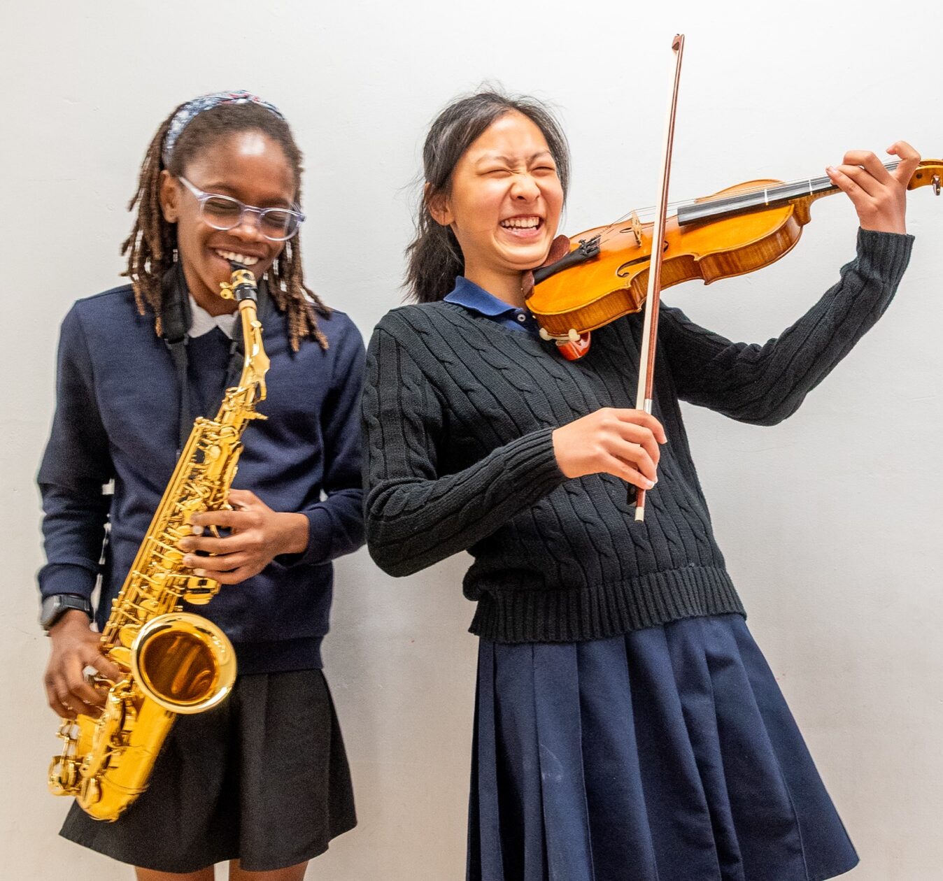 Two young girls in school uniforms play saxophone and violin together, smiling against a plain wall.