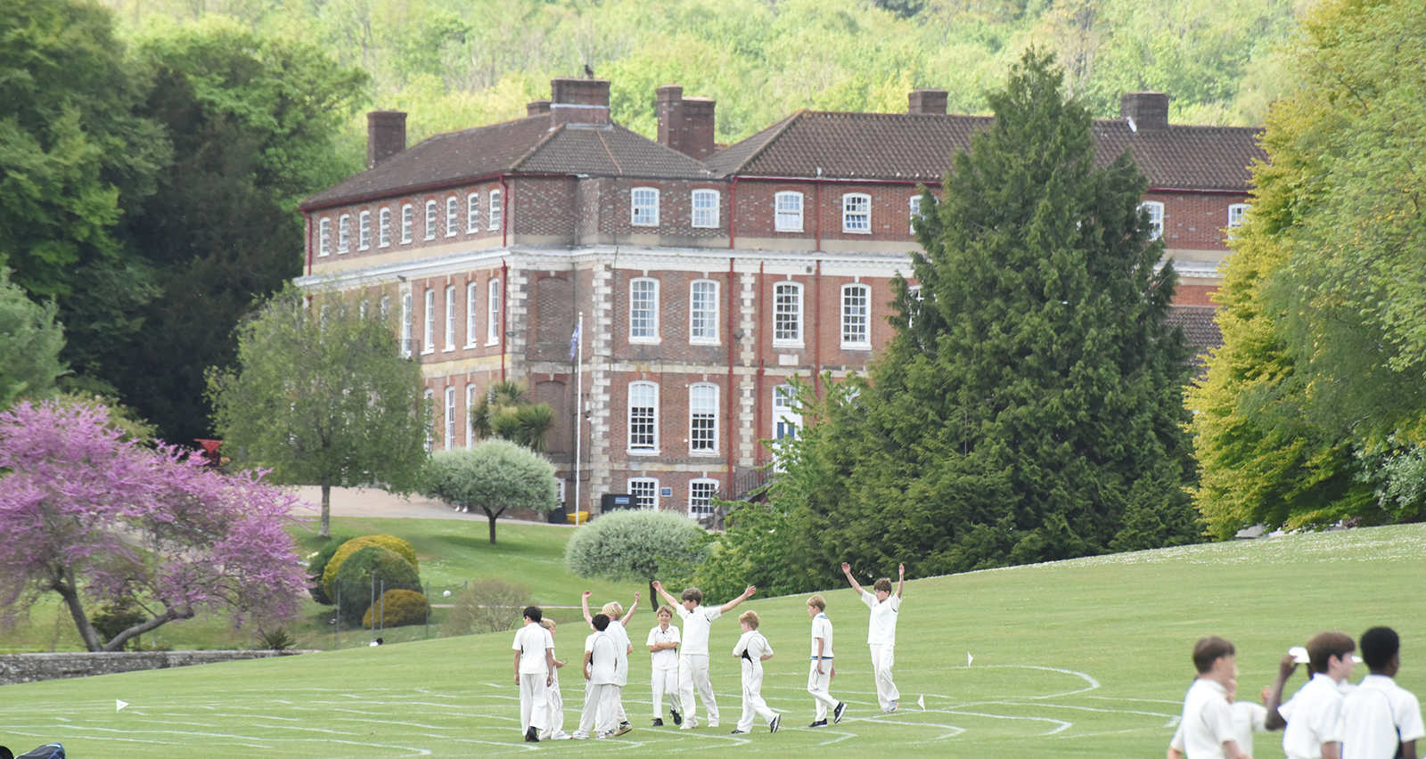 Group of students in white sports uniforms on a grassy field with a red-brick campus building and trees in the background.