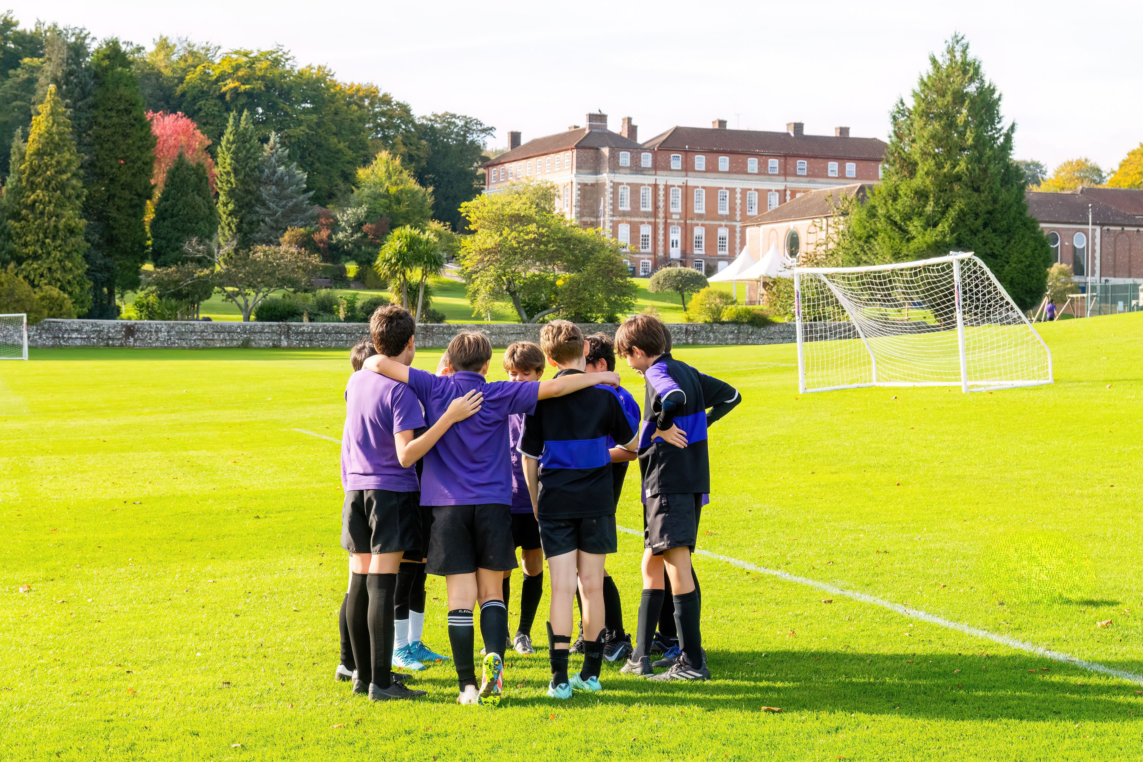 Group of young players in purple uniforms form a huddle on a sunny grassy soccer field, with a goal and a brick building in the background.
