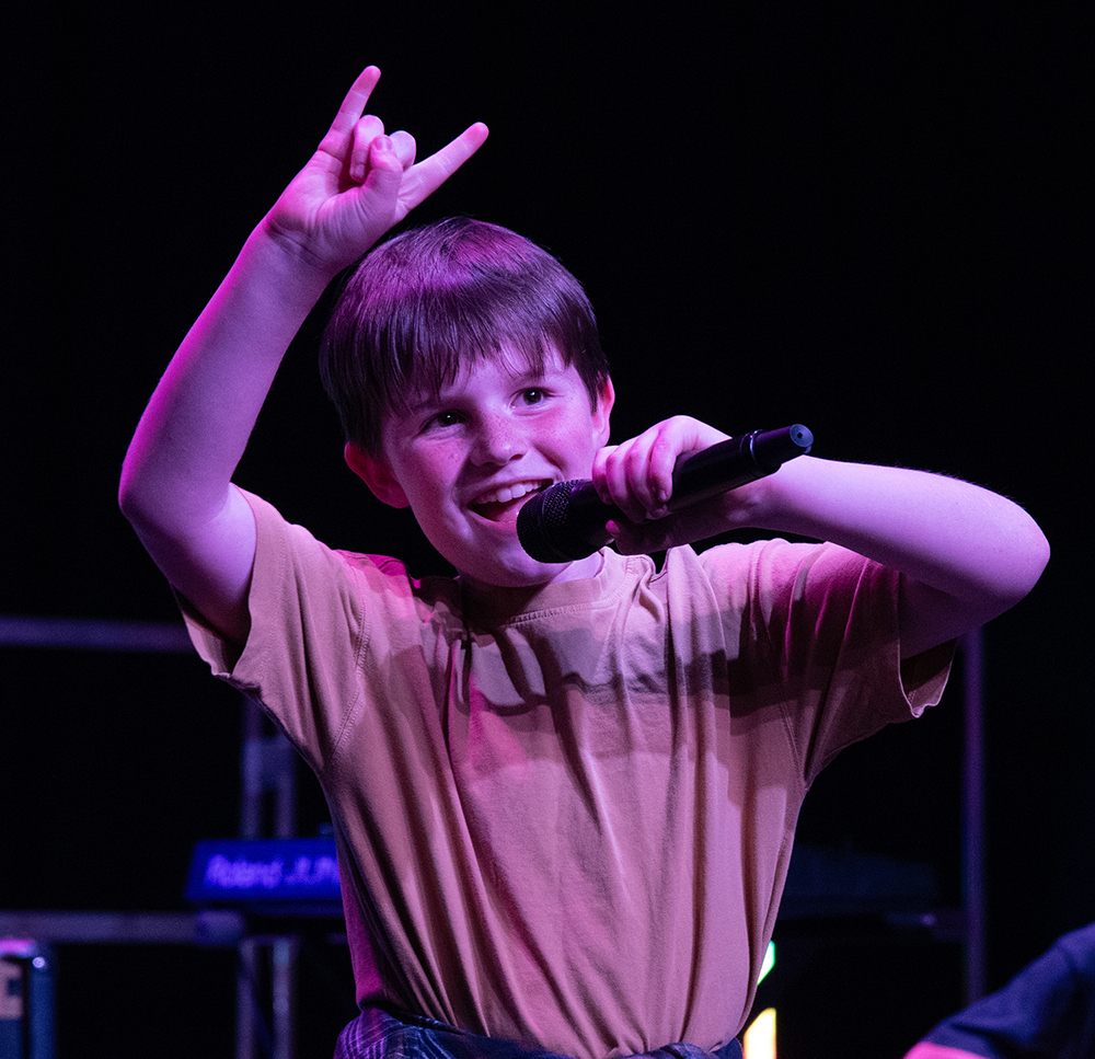 Boy performing on stage, singing into a microphone while flashing a 'rock on' gesture with his left hand under purple stage lighting.