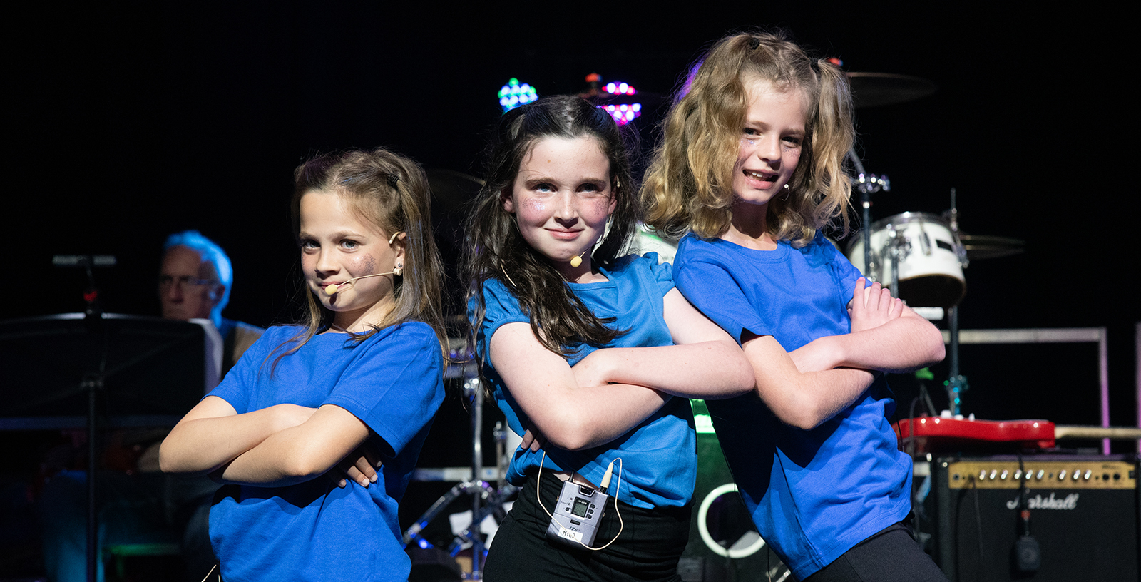 Three smiling girls in blue shirts pose with arms crossed on a stage during a performance, with band equipment in the background.