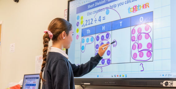 Student uses a stylus to solve a division math problem on a large interactive touchscreen in a classroom.