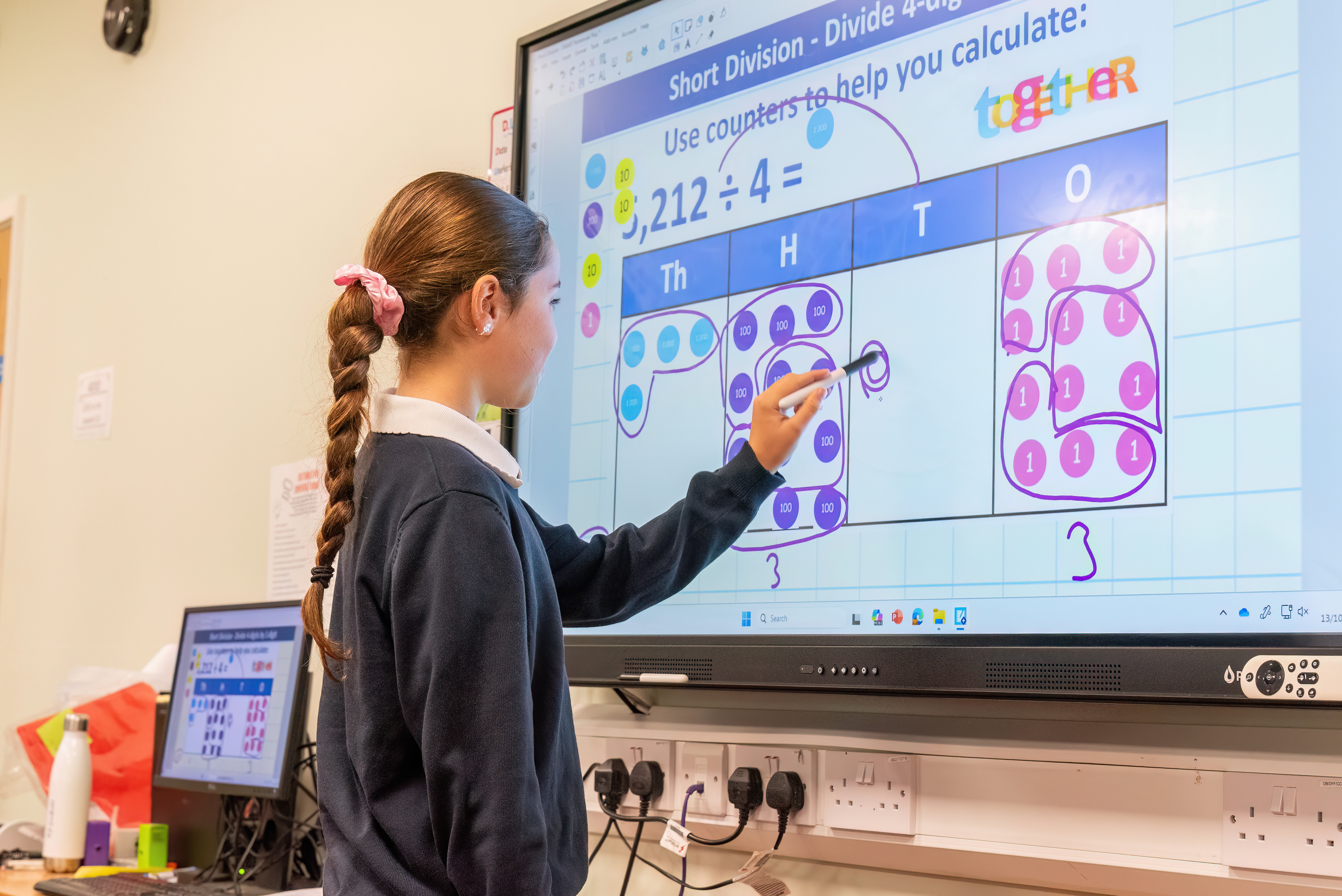 Student uses a stylus to solve a division math problem on a large interactive touchscreen in a classroom.