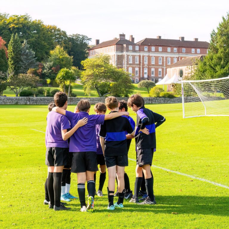 Group of young players in purple uniforms form a huddle on a sunny grassy soccer field, with a goal and a brick building in the background.