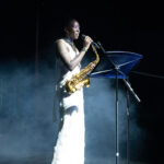 Female saxophonist in a white dress performs on a smoky stage, singing into a microphone beside a music stand.