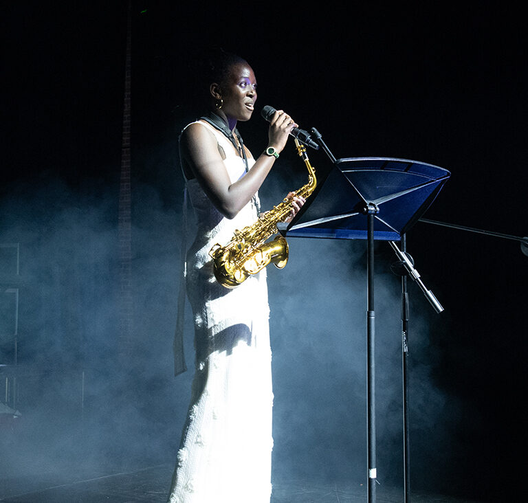Female saxophonist in a white dress performs on a smoky stage, singing into a microphone beside a music stand.