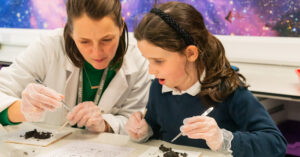 Two people in a lab coat and a student wearing gloves, using forceps to handle dark rock fragments at a lab bench with a colorful backdrop behind them.