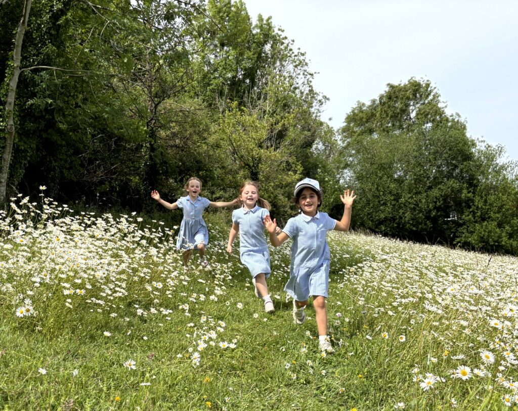 Three young girls in blue dresses run through a field of white daisies under a sunny sky.