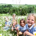Children smiling and kneeling in a field of white daisies with green hills in the background.